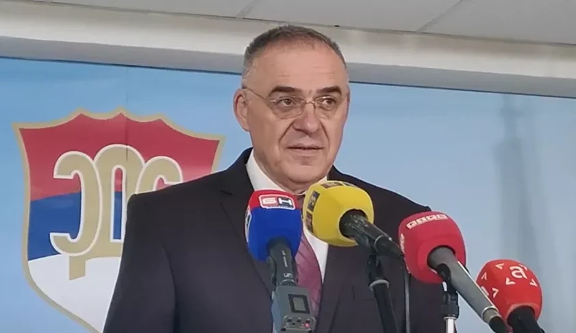 Muškarac u odijelu i naočarima govori ispred više mikrofona, sa grbom na kojem piše 'SNS' u pozadini.