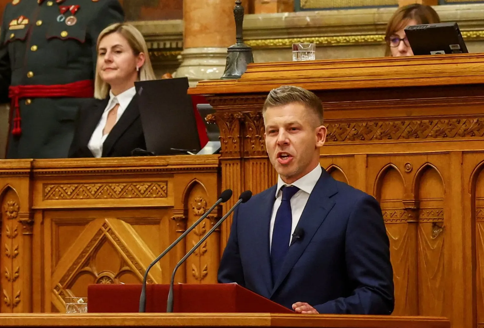 Političar u tamnom odijelu i plavoj kravati govori za govornicom ispred dva mikrofona, sa ženom u crnom sakou koja sjedi iza njega u parlamentarnoj sali.