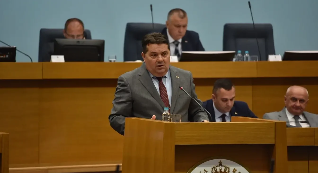Političar u odijelu drži govor za govornicom u skupštinskoj sali, ispred njega boca vode i čaša.
