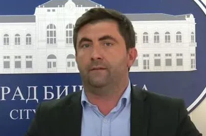 Političar, muškarac srednjih godina u crnom sakou i plavoj košulji, stoji ispred zgrade sa natpisom "GRAD BIJELIINA" i grbom grada.
