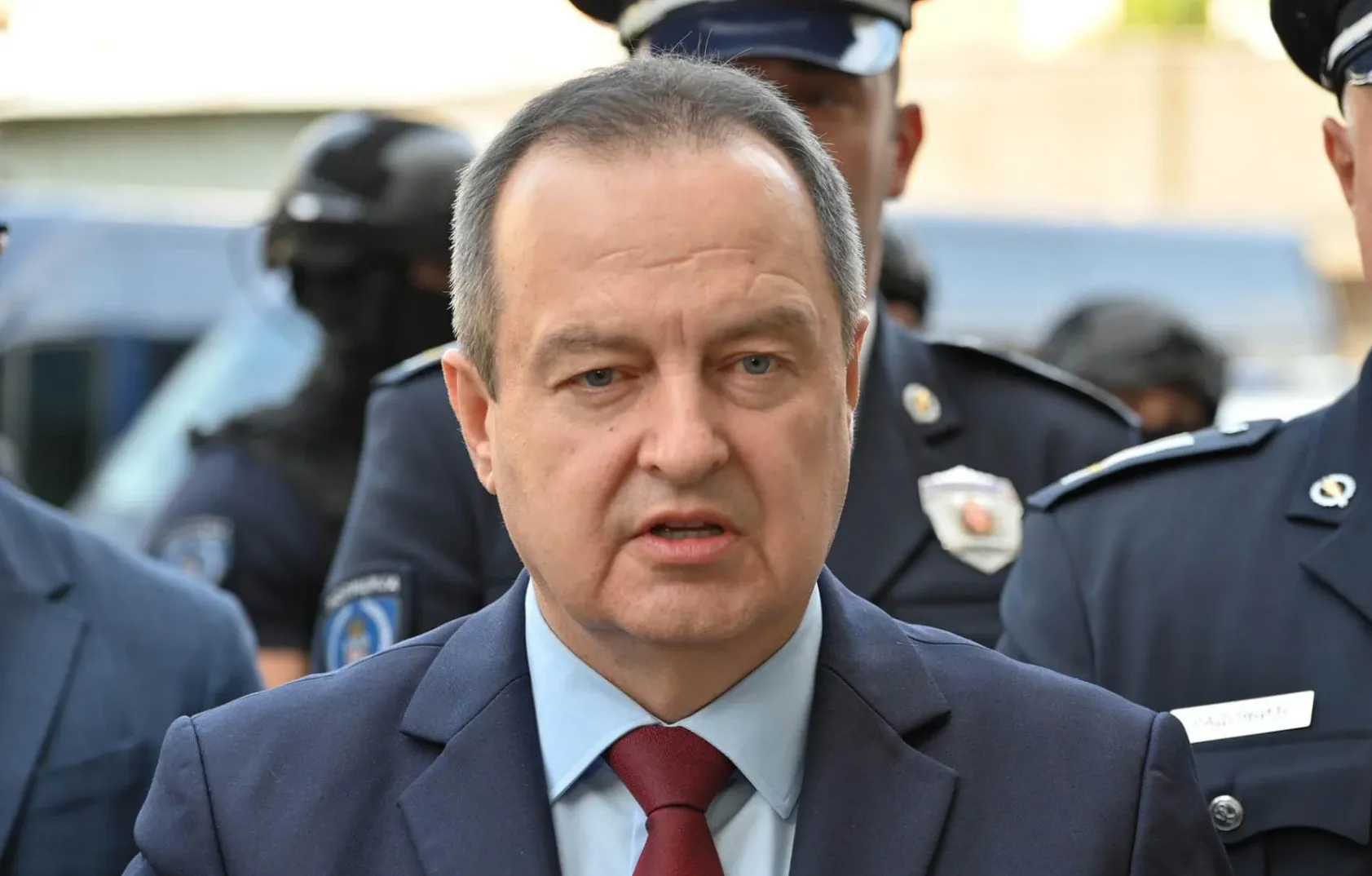 Političar u tamnom odijelu sa crvenom kravatom ispred grupe policajaca u uniformama.