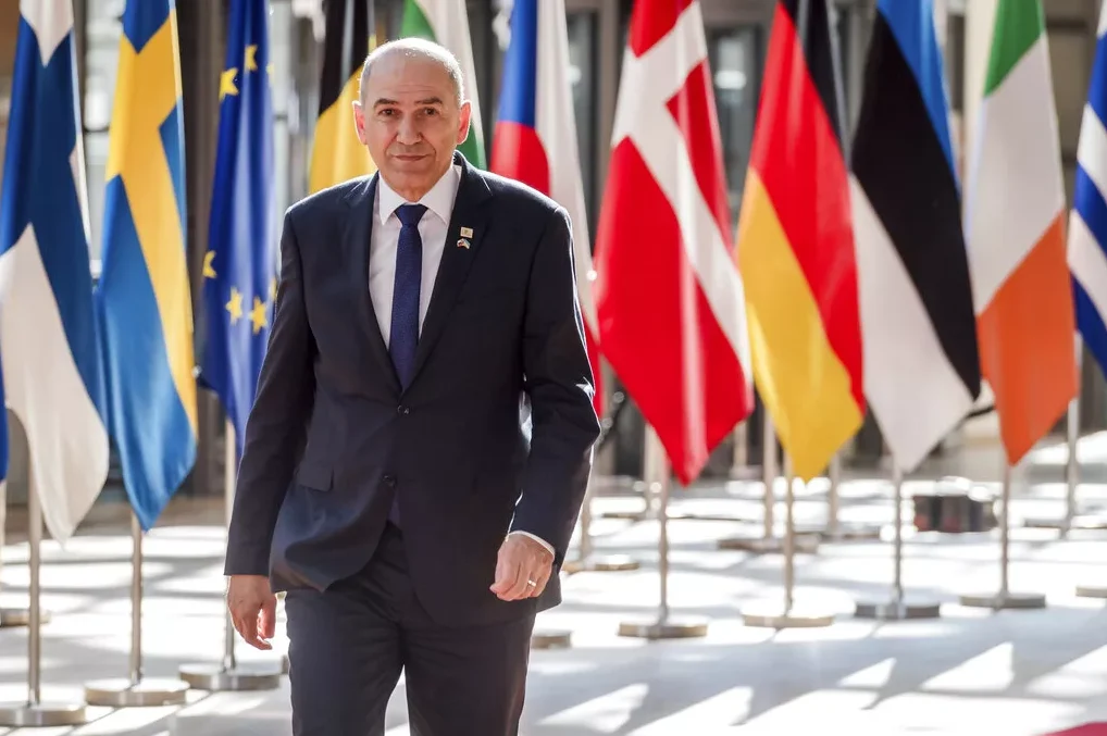 Političar u tamnom odijelu i plavoj kravati, stoji ispred niza zastava zemalja Evropske unije.