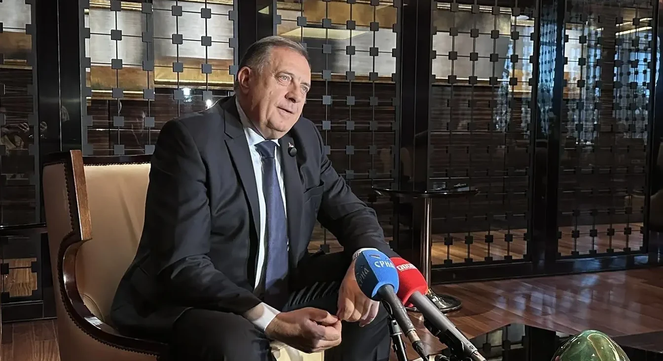 Političar u odijelu sjedi u stolici sa dva mikrofona ispred sebe.