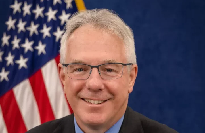 Portret muškarca srednjih godina sa sijedom kosom i naočarima, nasmiješenog lica, sa američkom zastavom u pozadini.
