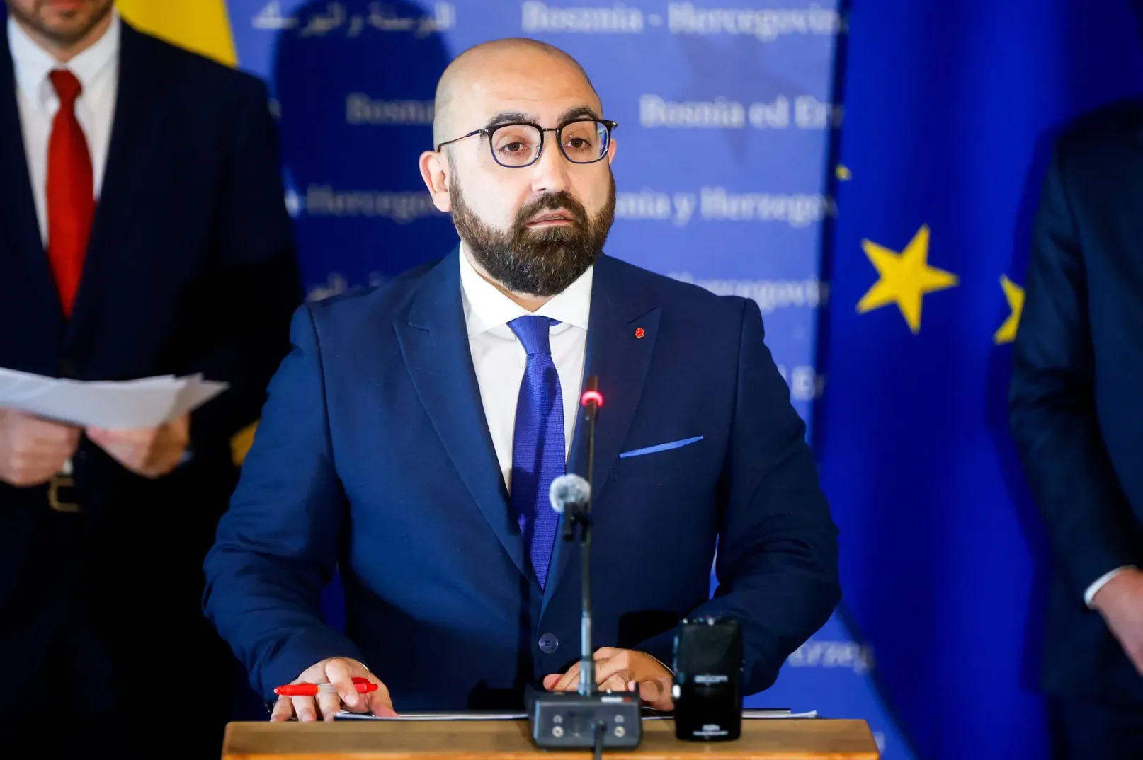 Političar sa naočarima i bradom u plavom odijelu sa plavom kravatom govori u mikrofon ispred zastava Bosne i Hercegovine i Evropske unije.