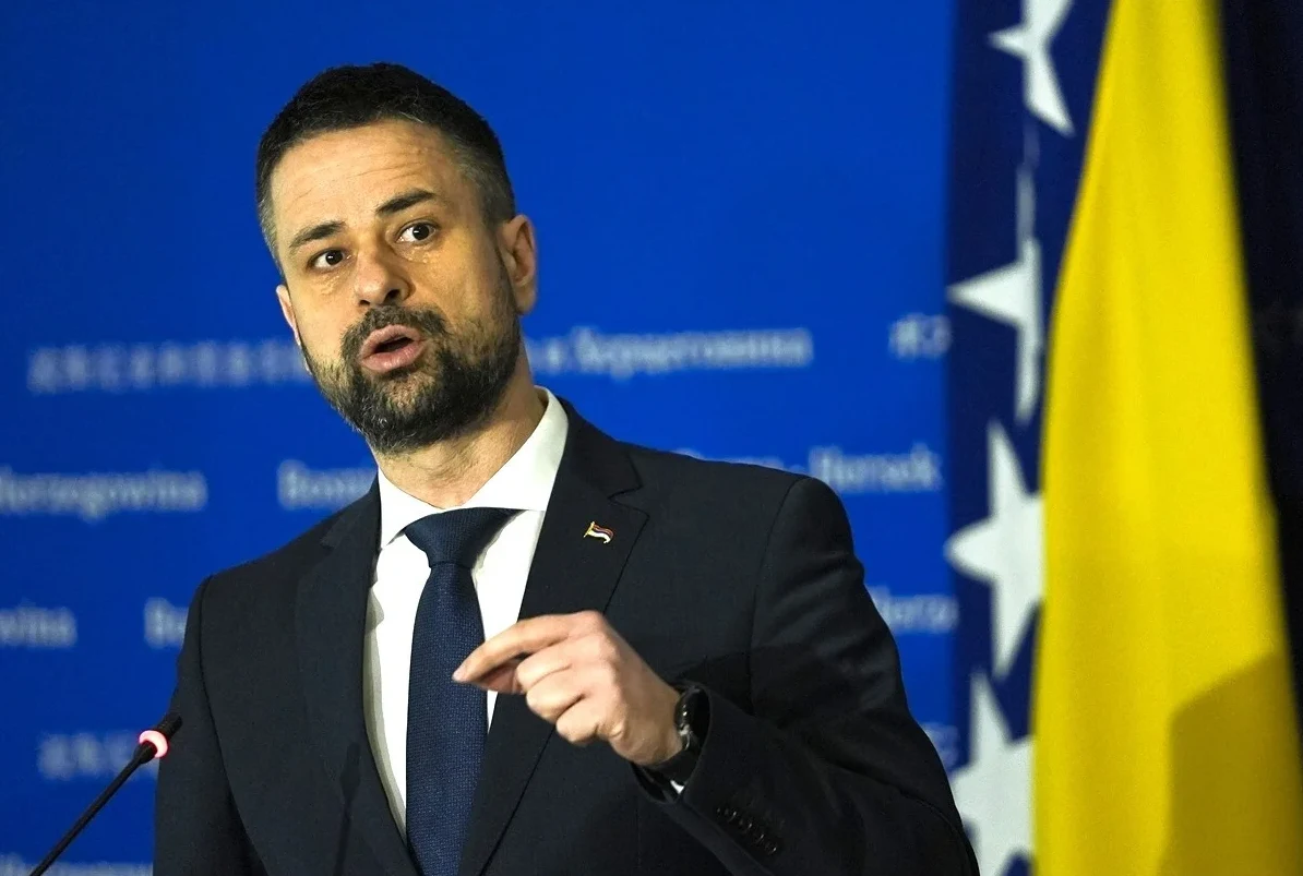 Političar u crnom odijelu i tamno plavoj kravati govori pred plavom pozadinom sa zastavom Bosne i Hercegovine.