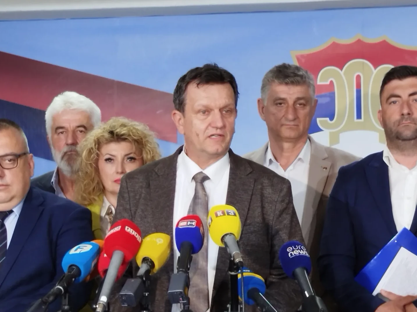 Grupacija političara okupljena oko govornice s brojnim mikrofonima