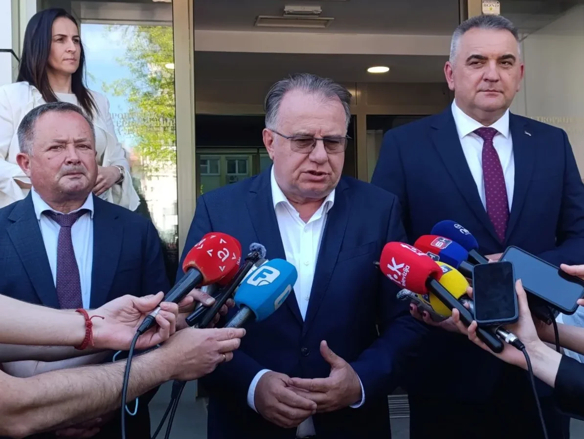 Političari u odijelima daju izjavu novinarima ispred zgrade, okruženi mikrofonima i telefonima za snimanje.