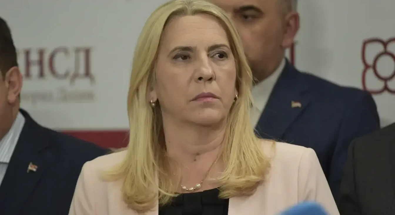 Žena sa plavom košuljom i zlatnim nakitom na vratu stoji ispred natpisa na ćirilici.