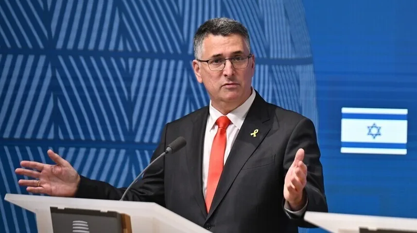 Portparol izraelske vlade u crnom odijelu i crvenoj kravati sa žutim simbolom na reveru, govori za govornicom ispred zastave Izraela.