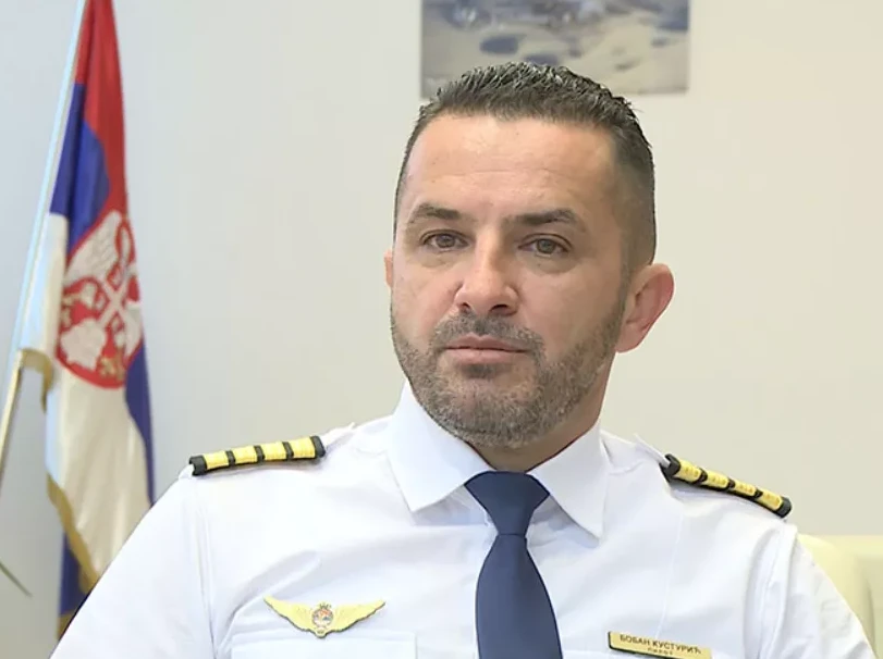 Portret Bojana Kusturice, oficira, u bijeloj uniformi sa tamno plavom kravatom, noseći amblem sa krilima i grbom Srbije na grudima. Na ramenima ima oznake činova. U pozadini se vidi zastava Srbije.