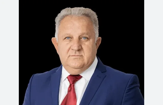 Portret starijeg muškarca sa sijedom kosom, plavim očima, u tamnoplavom odijelu i crvenoj kravati.