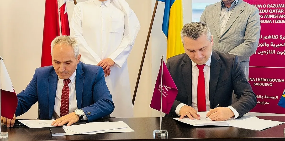 Dvojica muškaraca u odijelima potpisuju dokumente na sastanku, uz zastave Katara i Bosne i Hercegovine.