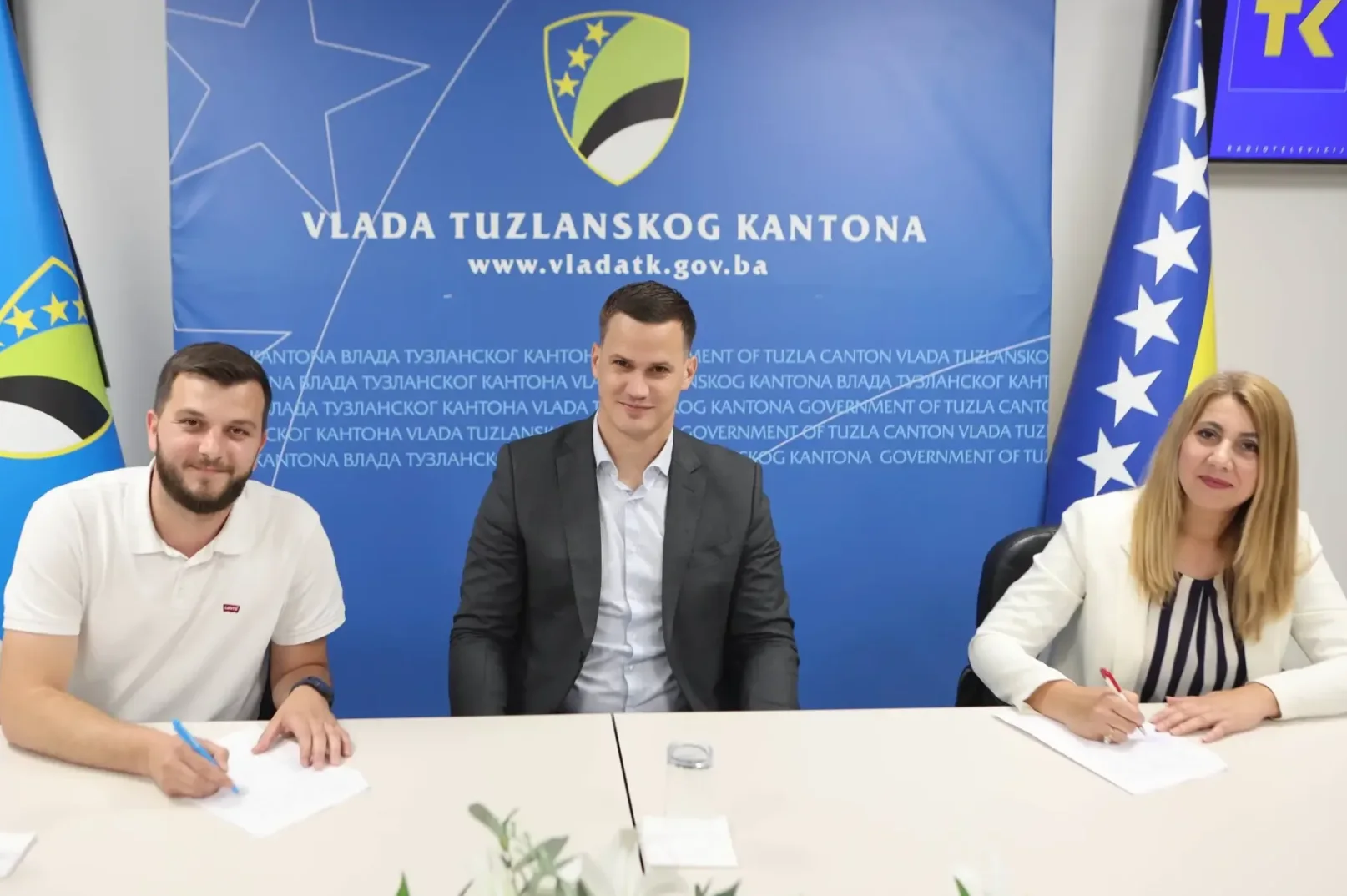 Tri osobe sjede za stolom ispred natpisa "Vlada Tuzlanskog kantona", dvoje potpisuju dokumente.