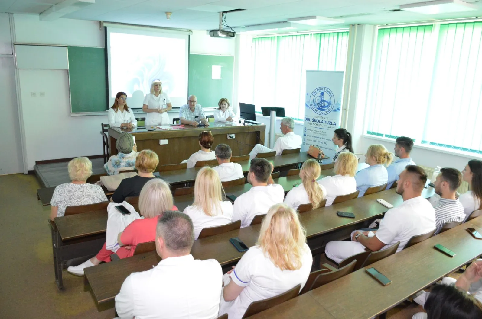 Grupa ljudi sjedi u učionici slušajući predavanje; na prednjoj strani, troje predavača stoje iza podijuma, a jedan od njih drži prezentaciju na projekcionom platnu.