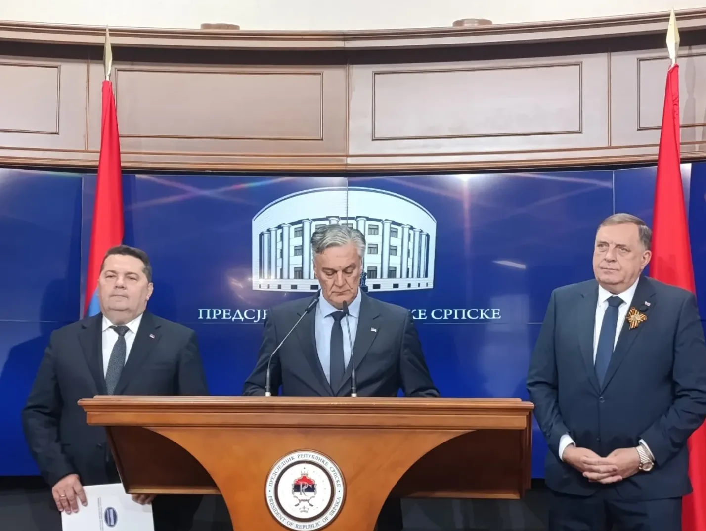 Tri muškarca u tamnim odijelima stoje ispred postolja za govore sa zastavama Republike Srpske u pozadini. Jedan muškarac govori za govornicu, dok druga dvojica stoje pored njega, gledajući prema publici.