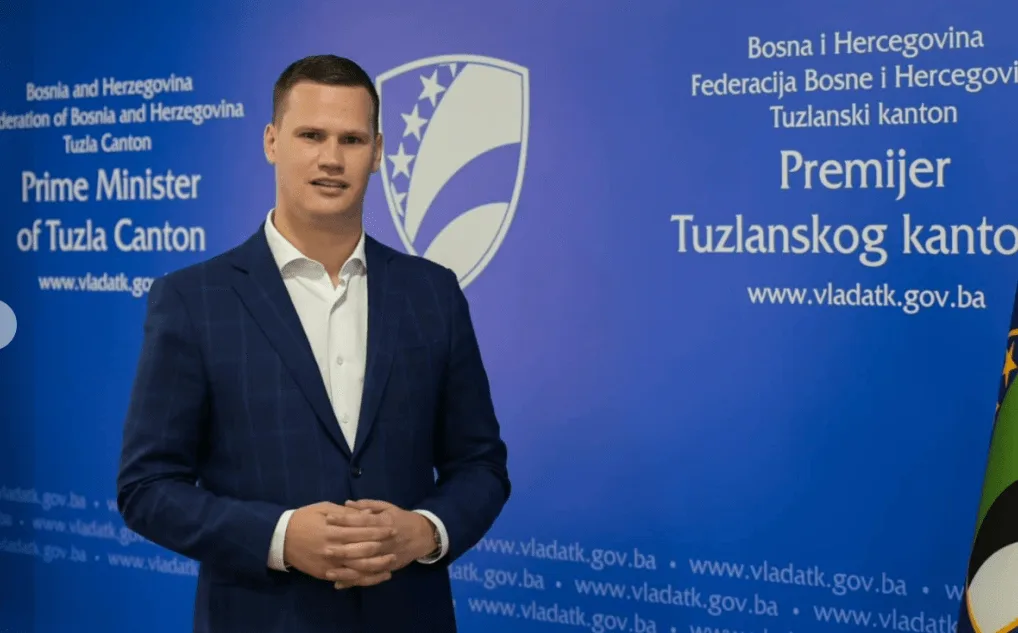 Premijer Tuzlanskog kantona u tamnoplavom odijelu sa bijelom košuljom, stoji ispred plave pozadine sa logom kantona i natpisom na bosanskom jeziku.