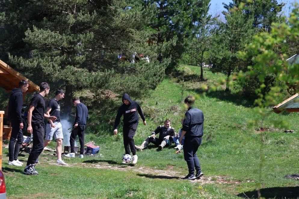 Grupa mladića igra fudbal u prirodi dok se drugi odmaraju i roštiljaju.