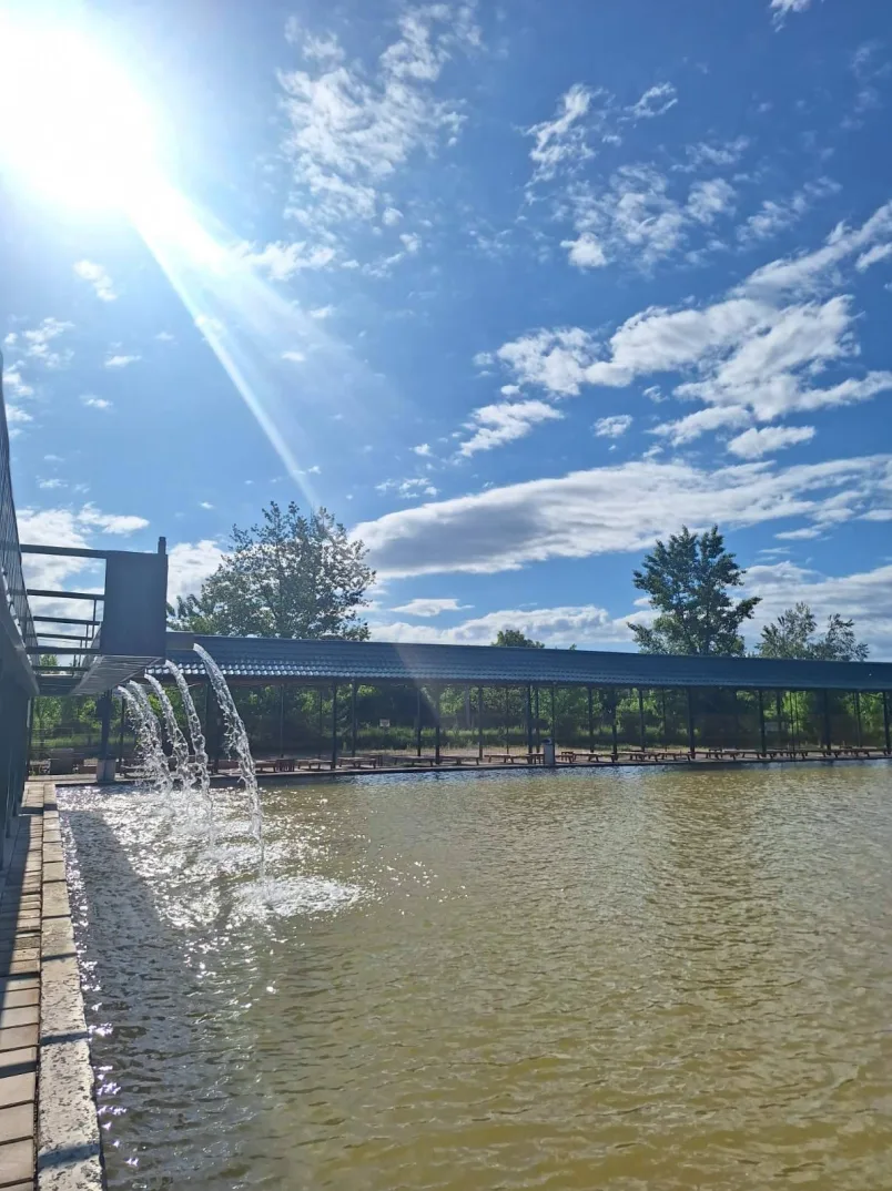 Ribnjak sa više fontana koje se ulivaju u vodu, izgrađena je duga nadstrešnica sa stolovima i klupama za sjedenje, te je nebo plavo sa bijelim oblacima.