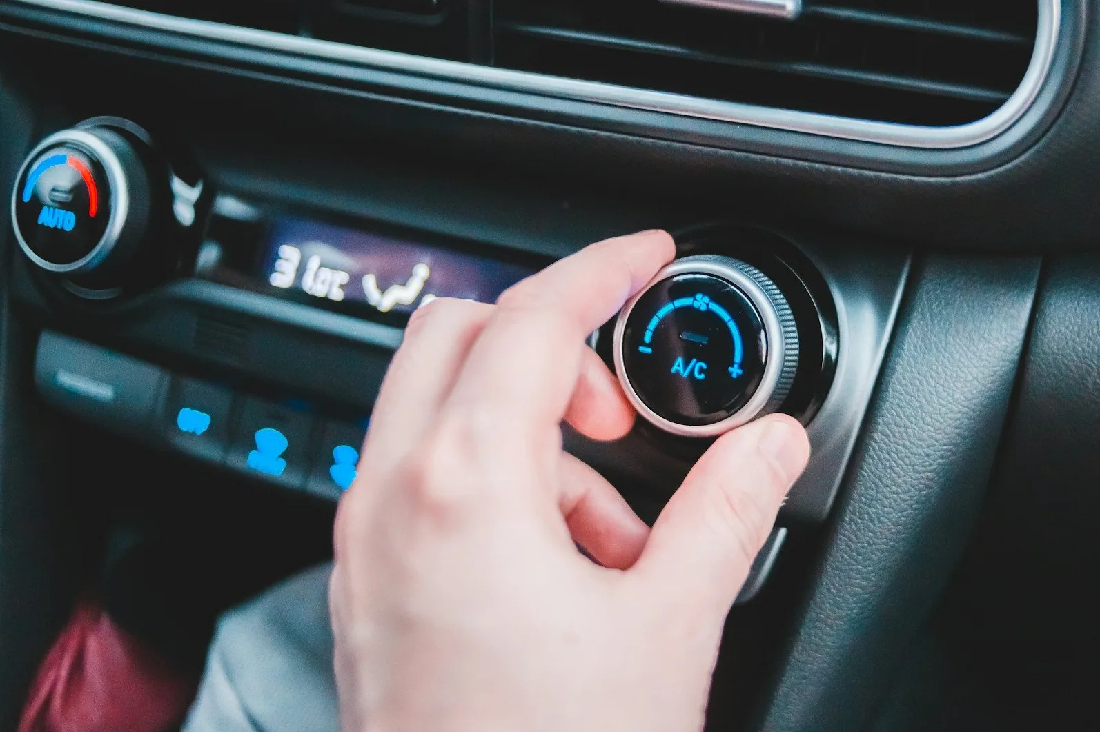 Bliski prikaz ruke koja okreće točkić za podešavanje klime u automobilu, sa osvijetljenim simbolom A/C.