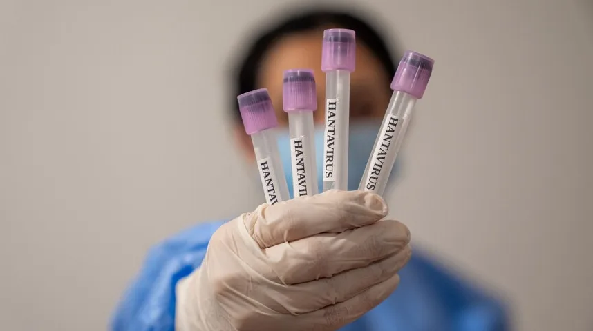 Ruke u bijelim rukavicama drže nekoliko epruveta sa ljubičastim čepovima na kojima je ispisano 'HANTAVIRUS'. Osoba nosi plavi zaštitni mantil.