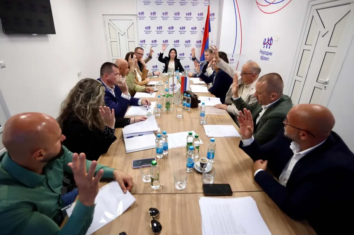 Grupa ljudi sjedi za stolom na sastanku, svi sa podignutim rukama kao da glasaju.