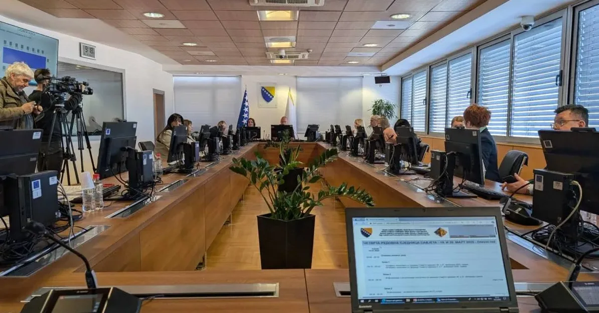 Ljudi sjede za dugačkim stolom u konferencijskoj sali, okruženi računalima i monitorima. Na stolu ispred njih nalaze se boce vode i papiri. Na sredini stola je postavljena sobna biljka u crnoj saksiji. Zastava Bosne i Hercegovine i bijela zastava su istaknute na zidu, dok na drugoj strani prostorije prozori imaju zatamnjene roletne. U prednjem planu vidljivi su laptop sa ispisanim dokumentom i mikrofonom.