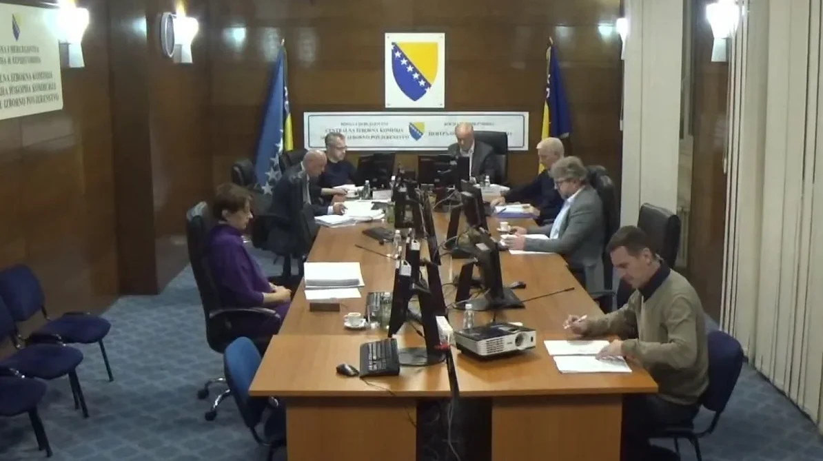Grupa ljudi sjedi za dugim drvenim stolom u prostoriji koja izgleda kao sala za sastanke. Oni rade na papirima, gledaju u monitore i komuniciraju jedni s drugima. Na stolu su, pored radnih materijala, i dva projektora, kao i službena zastava Bosne i Hercegovine.