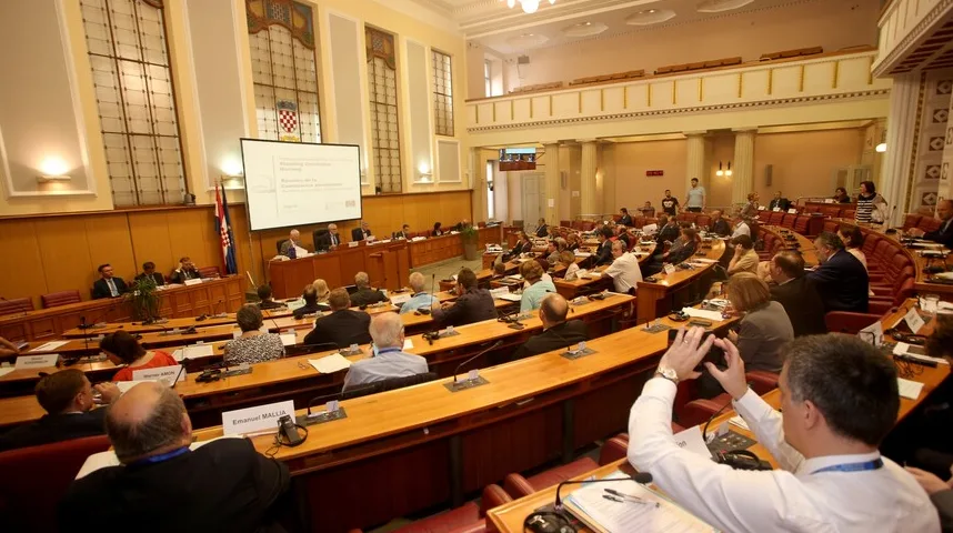 Pogled na salu za sastanke u kojoj sjede ljudi za drvenim stolovima, s desne strane neko slika telefon.
