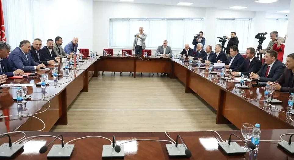 Grupa muškaraca u odijelima sjedi za velikim konferencijskim stolom u moderno opremljenoj sali, dok jedan čovjek snima mobitelom u ruci.