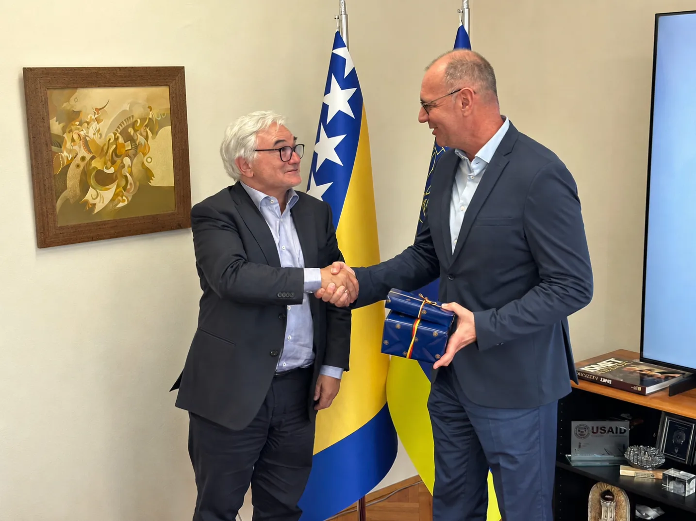 Dva muškarca u odijelima se rukuju ispred zastave Bosne i Hercegovine, jedan muškarac predaje poklon drugome.