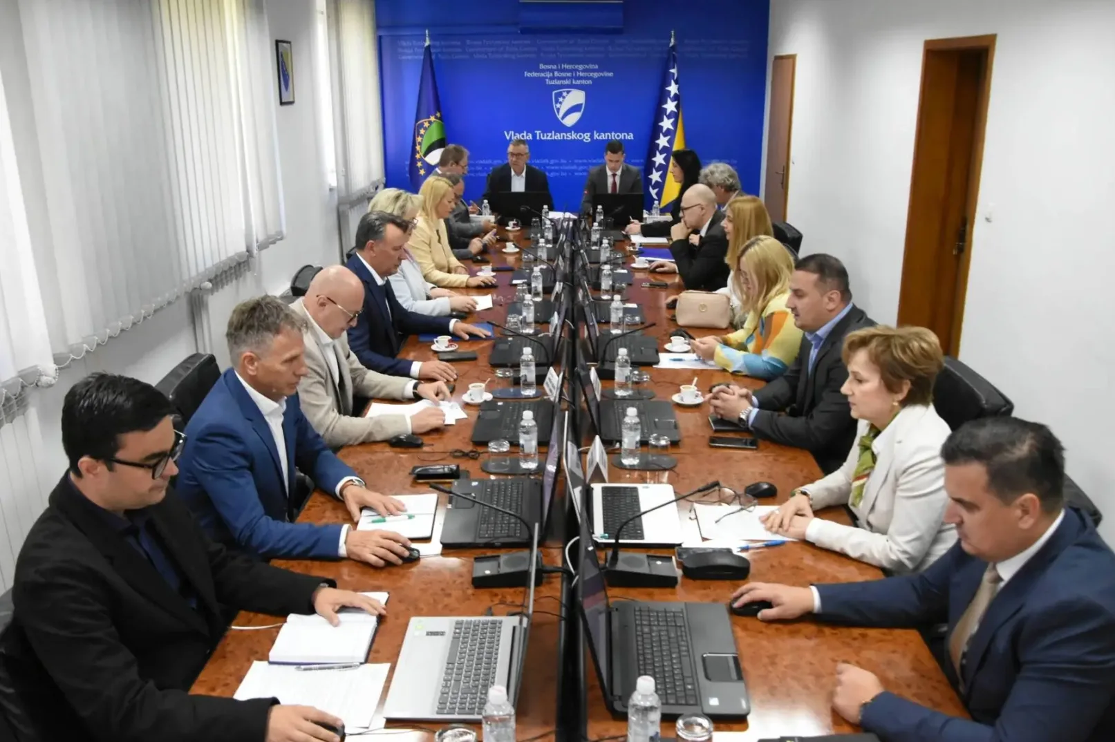 Grupa ljudi sjedi za stolom na sastanku u prostoriji sa plavim zidovima i zastavama Bosne i Hercegovine i Tuzlanskog kantona.
