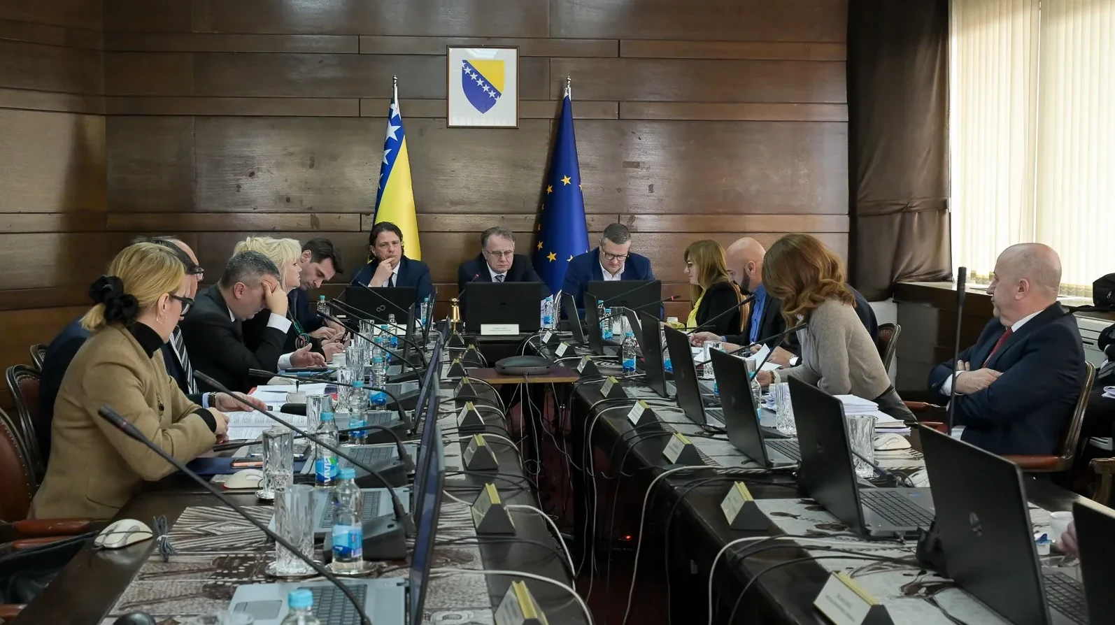 Zvaničnici sjede za dugačkim stolom u sali, okruženi laptopima i dokumentima.