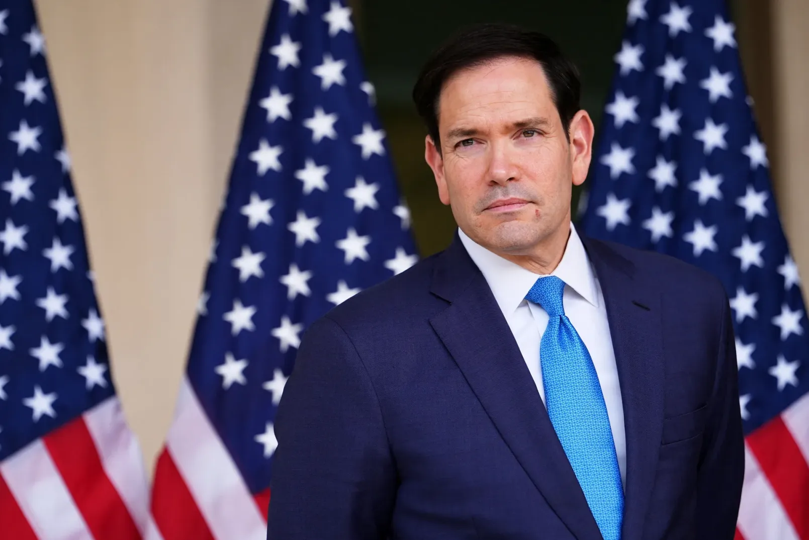 Senator Marco Rubio u tamnoplavom odijelu i plavoj kravati, sa tri američke zastave u pozadini.