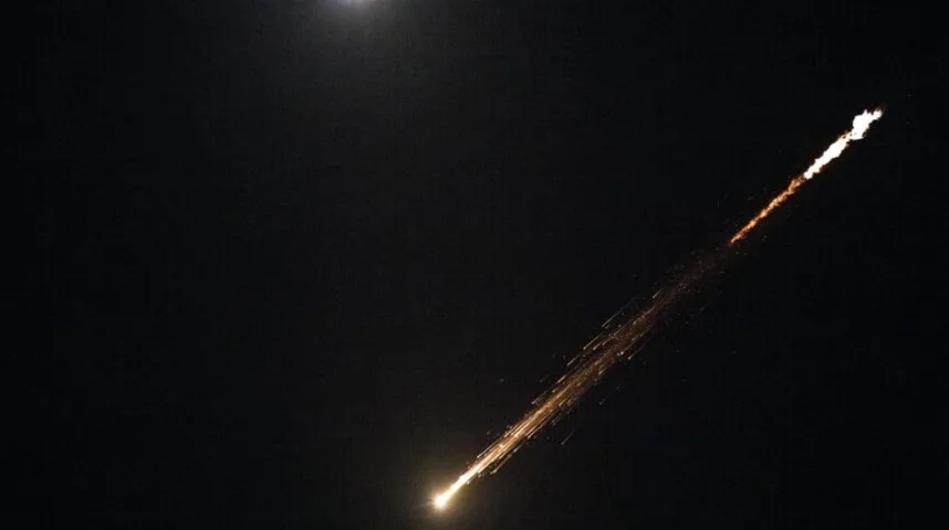 Svijetli meteor ili zvijezda padalica ostavlja narandžasti i žuti trag na tamnom noćnom nebu, dok se mjesečina nazire u gornjem lijevom uglu.