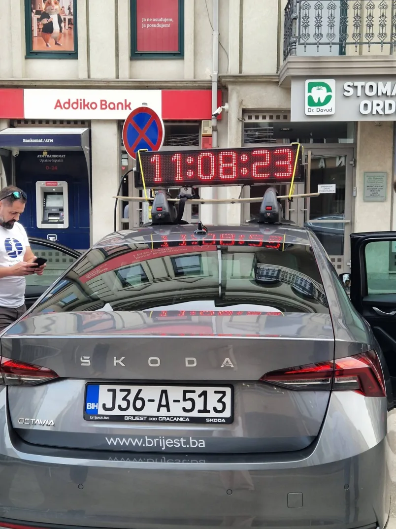 Siva Škoda Octavia sa digitalnim displejem na krovu koji prikazuje vrijeme 11:08:23, stoji ispred Addiko Banke i stomatološke ordinacije.
