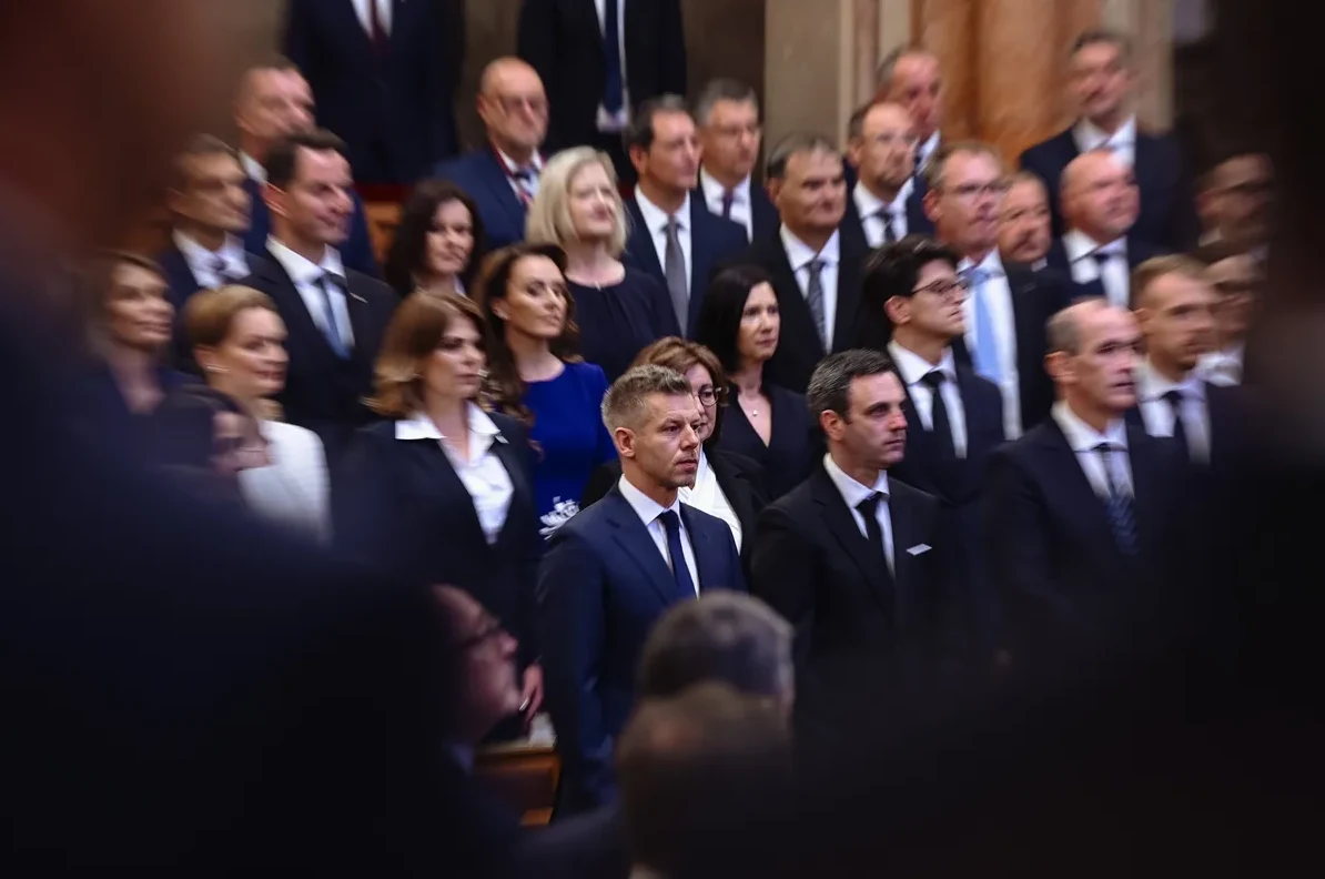 Grupa ljudi u formalnoj odjeći, vjerovatno političara ili zvaničnika, kako stoje ili sjede u sali.