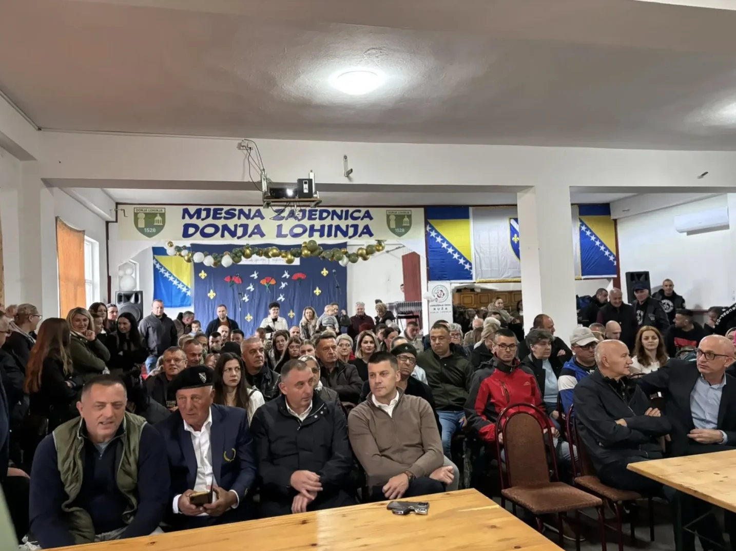 Skup ljudi u sali, ispred njih zastave Bosne i Hercegovine i transparent "Mjesna zajednica Donja Lohinja"