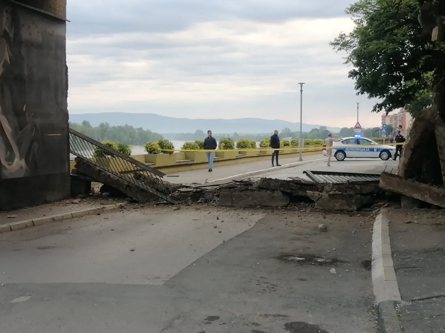 Slika prikazuje urušeni most sa betonskim krhotinama na cesti, uz prisustvo policijskog vozila i nekoliko osoba.