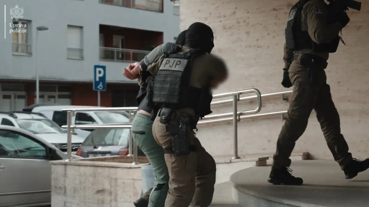 Pripadnici specijalne jedinice policije u opremi, s fantomkama na licima, vrše lišenje slobode osobe na stepeništu ispred zgrade. Jedan policajac drži uhapšenog, dok drugi, naoružan, ide iza njih.