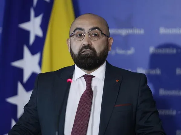 Muškarac u tamnom odijelu i tamno crvenoj kravati, sa naočalama i bradom, stoji ispred zastave Bosne i Hercegovine.