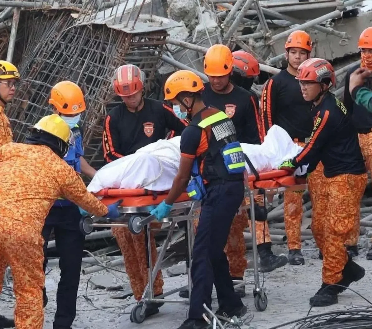 Tim spasioca u narandžastim uniformama i zaštitnim kacigama nosi nosila s osobom umotanom u bijelo platno, okruženi ruševinama i metalnim konstrukcijama.