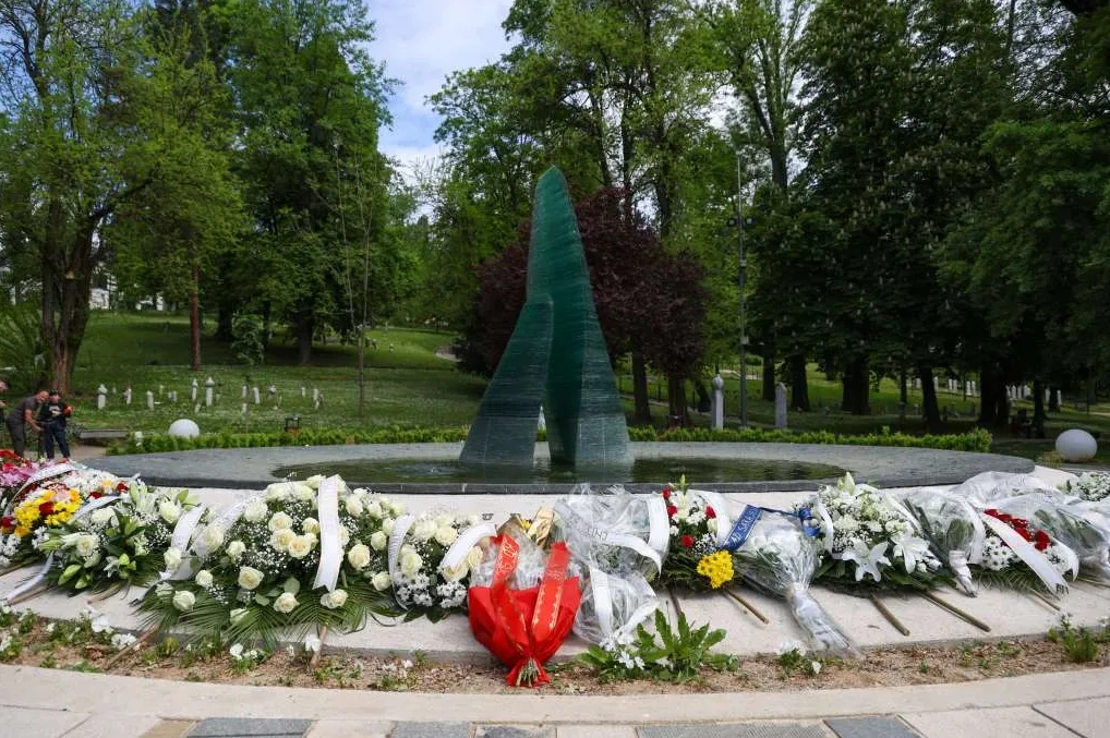 Brojni vijenci cvijeća položeni su ispred modernog staklenog spomenika u parku prepunom zelenila i bijelih nadgrobnih ploča.
