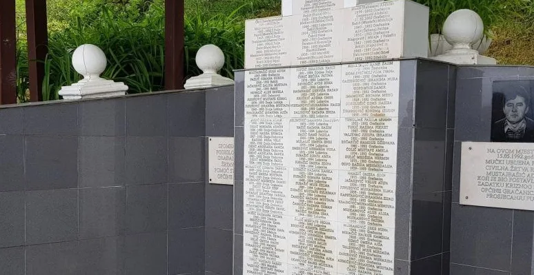 Spomen-obilježje sa uklesanim imenima žrtava rata na crnoj mermernoj ploči, uz bijele stubove i zelenilo u pozadini.