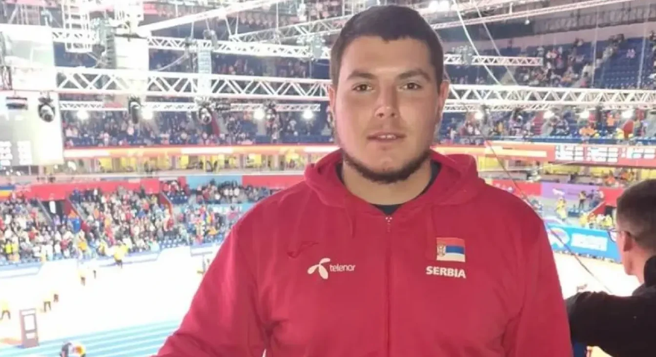 Mladić u crvenoj dukserici sa natpisom "SERBIA" i zastavom Srbije na lijevoj strani grudi, snimljen je na tribinama sportskog događaja. U pozadini se vide gledaoci i sportska dvorana sa osvjetljenjem.
