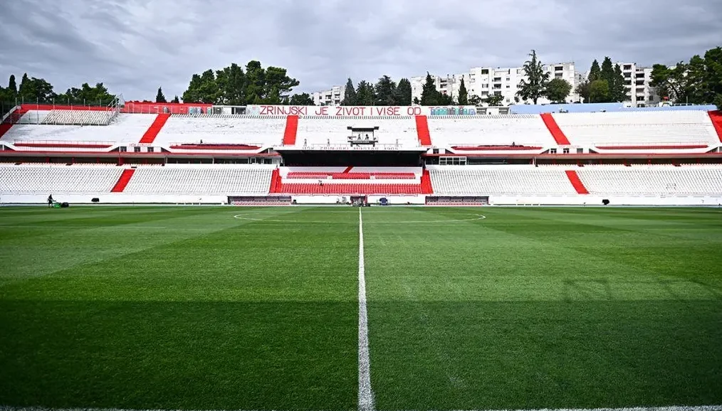Fudbalski stadion sa zelenim terenom i bijelim sjedalicama sa crvenim detaljima, na tribinama natpis "ZRINJSKI JE ŽIVOT I VIŠE OD TOGA"