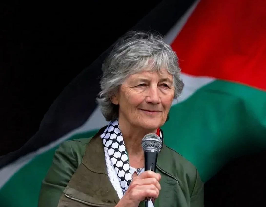 Žena sa sijedom kosom i palestinskom maramom govori u mikrofon ispred palestinske zastave
