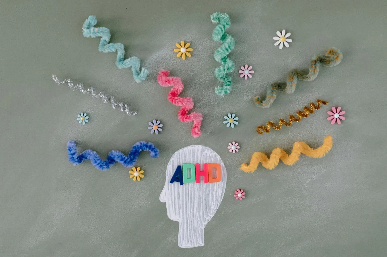 Bijela stilizovana silueta glave sa slovima 'ADHD' napisanima šarenim slovima unutar glave, okružena raznobojnim uvijenim nitima i malim cvjetićima.