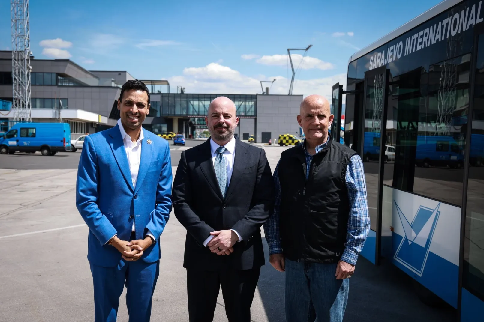 Tri muškarca stoje ispred zgrade Međunarodnog aerodroma Sarajevo pored autobusa sa natpisom "SARAJEVO INTERNATIONAL".