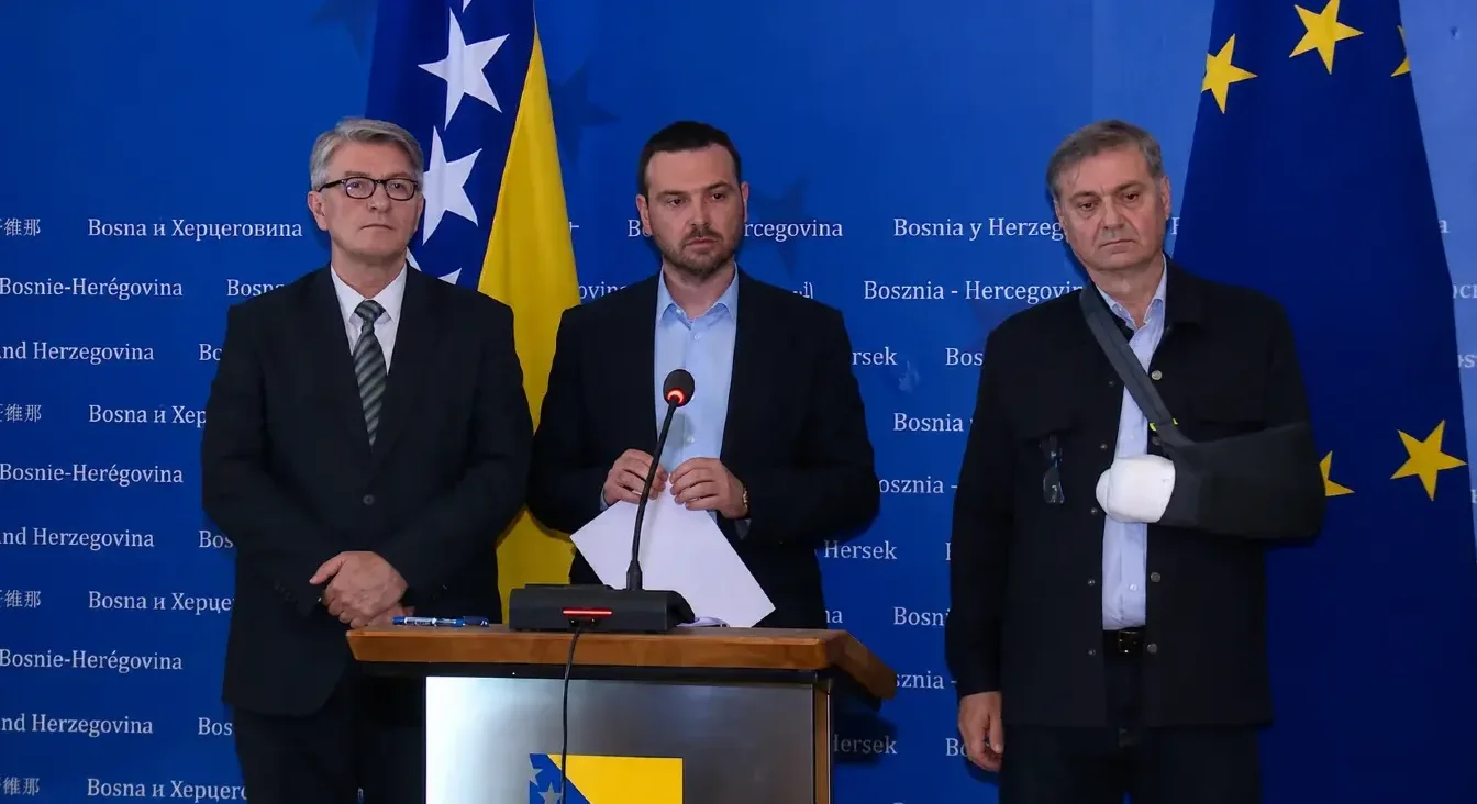 Tri muškarca stoje ispred zastave Bosne i Hercegovine i Evropske unije, jedan od njih nosi ortopedsku ortozu na ruci.
